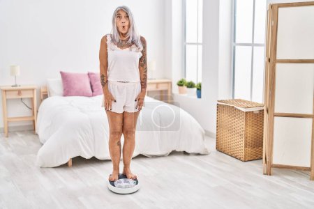 Photo for Middle age woman with grey hair standing on weight machine at the bedroom scared and amazed with open mouth for surprise, disbelief face - Royalty Free Image