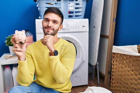 Photo for Hispanic man doing laundry holding piggy bank serious face thinking about question with hand on chin, thoughtful about confusing idea - Royalty Free Image