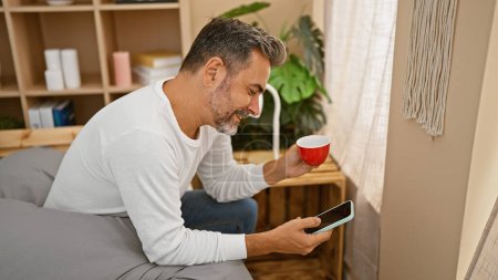 Téléchargez les photos : Jeune homme hispanique aux cheveux gris utilisant un smartphone buvant du café assis sur le lit dans la chambre - en image libre de droit
