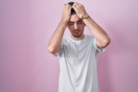 Foto de Young hispanic man standing over pink background suffering from headache desperate and stressed because pain and migraine. hands on head. - Imagen libre de derechos