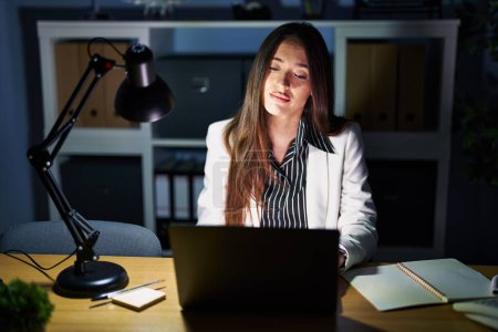 Téléchargez les photos : Jeune femme brune travaillant au bureau la nuit avec un ordinateur portable à la recherche somnolent et fatigué, épuisé par la fatigue et la gueule de bois, les yeux paresseux le matin. - en image libre de droit
