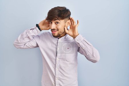 Téléchargez les photos : Homme arabe avec la barbe debout sur fond bleu essayant d'entendre les deux mains sur le geste de l'oreille, curieux de commérages. problème auditif, sourd - en image libre de droit