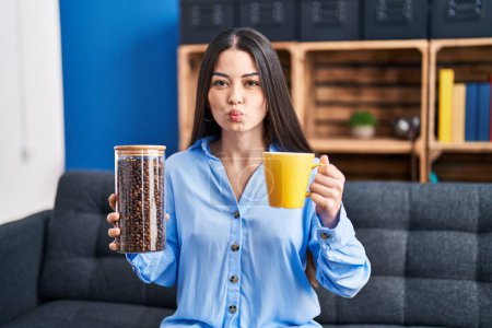 Photo for Young brunette woman holding coffee beans and cup of coffee looking at the camera blowing a kiss being lovely and sexy. love expression. - Royalty Free Image