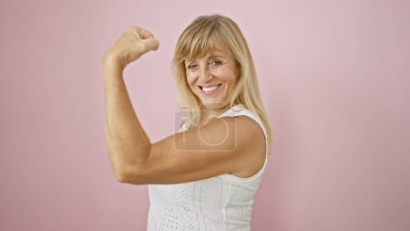Die schöne blonde Frau mittleren Alters strahlt vor Selbstvertrauen und Freude und beugt ihren starken Arm über einen isolierten rosafarbenen Hintergrund. Ausdruck von Glück, sportlich reifer Erwachsener genießt lustige Pose.