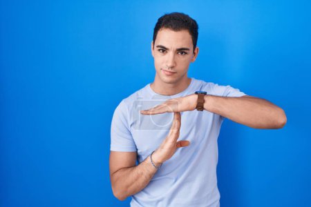 Foto de Young hispanic man standing over blue background doing time out gesture with hands, frustrated and serious face - Imagen libre de derechos
