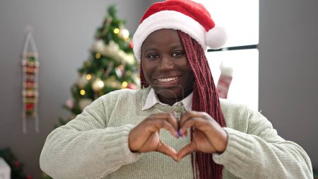 Téléchargez les photos : Femme africaine avec des cheveux tressés souriant par arbre de Noël faisant forme de coeur avec les mains à la maison - en image libre de droit