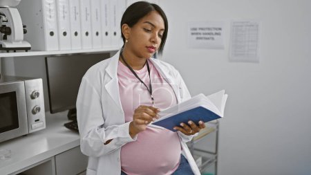 Téléchargez les photos : Jeune femme scientifique enceinte, immergée dans un livre de chimie profonde lu à son poste de travail, fusionnant maternité et recherche médicale. - en image libre de droit