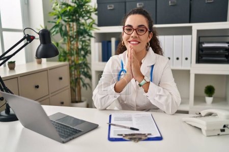 Photo for Young hispanic woman wearing doctor uniform and stethoscope praying with hands together asking for forgiveness smiling confident. - Royalty Free Image
