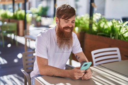 Téléchargez les photos : Jeune rousse homme en utilisant smartphone assis sur la table à la terrasse du café - en image libre de droit