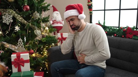 Foto de Joven hispano sufriendo por dolor de cabeza y estómago sentado junto al árbol de Navidad en casa - Imagen libre de derechos