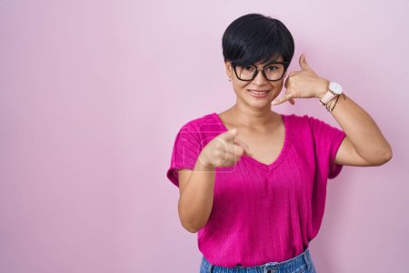 Téléchargez les photos : Jeune femme asiatique aux cheveux courts debout sur fond rose souriant faisant parler au téléphone geste et vous pointant du doigt. Appelle-moi. - en image libre de droit