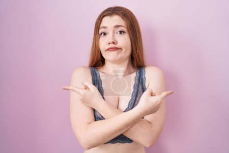 Photo for Redhead woman wearing lingerie over pink background pointing to both sides with fingers, different direction disagree - Royalty Free Image