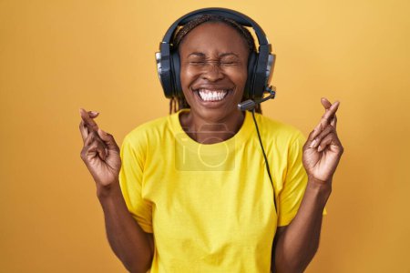 Téléchargez les photos : Femme afro-américaine écoutant de la musique à l'aide d'écouteurs geste doigt croisé souriant avec espoir et les yeux fermés. chance et concept superstitieux. - en image libre de droit