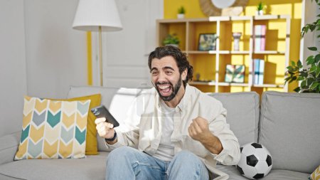 Photo for Young hispanic man watching soccer match using smartphone celebrating at home - Royalty Free Image