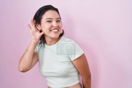 Téléchargez les photos : Jeune femme hispanique debout sur fond rose souriant avec la main sur l'oreille en écoutant une rumeur ou des commérages. concept de surdité. - en image libre de droit