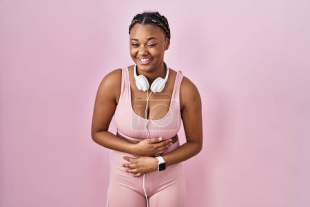 Téléchargez les photos : Femme afro-américaine avec des tresses portant des vêtements de sport et des écouteurs souriant et riant fort à haute voix parce que drôle blague folle avec les mains sur le corps. - en image libre de droit