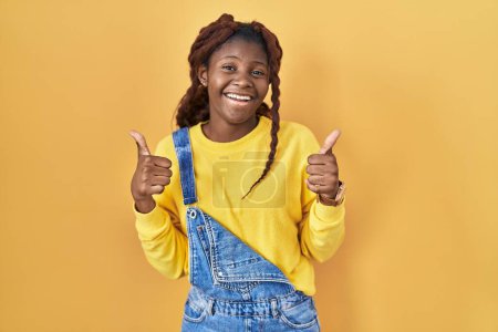 Téléchargez les photos : Femme africaine debout sur fond jaune signe de succès faisant geste positif avec la main, pouces levés souriant et heureux. expression joyeuse et geste gagnant. - en image libre de droit