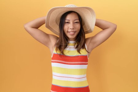 Foto de Mujer china de mediana edad con sombrero de verano sobre fondo amarillo relajante y estiramiento, brazos y manos detrás de la cabeza y el cuello sonriendo feliz - Imagen libre de derechos