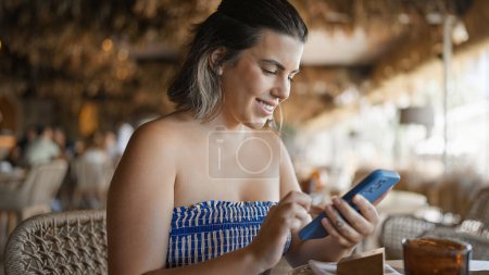 Photo for Young hispanic woman smiling happy using smartphone sitting on the table at the restaurant - Royalty Free Image