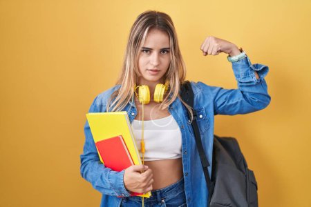 Foto de Mujer rubia joven usando mochila de estudiante y sosteniendo libros persona fuerte mostrando el músculo del brazo, confiado y orgulloso de poder - Imagen libre de derechos