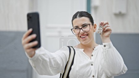 Photo for Young beautiful hispanic woman taking a picture showing keys at new house front door - Royalty Free Image