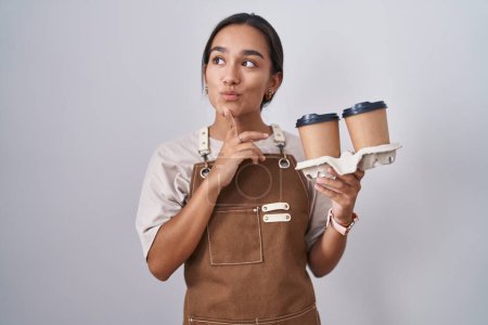 Foto de Mujer hispana joven usando delantal de camarera profesional sosteniendo café pensando concentrado en la duda con el dedo en la barbilla y mirando hacia arriba preguntándose - Imagen libre de derechos
