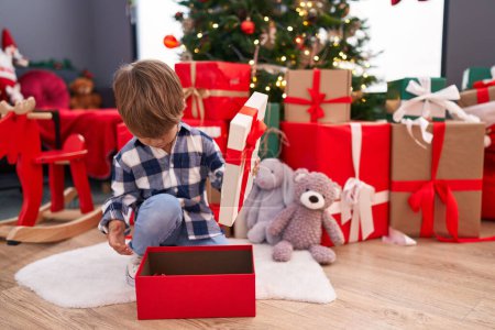 Téléchargez les photos : Adorable garçon hispanique déballer cadeau assis par arbre de Noël à la maison - en image libre de droit