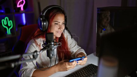 Téléchargez les photos : Vibrant jeune rousse femme streamer jouant joyeusement jeu virtuel sur smartphone dans la salle de jeu sombre - en image libre de droit
