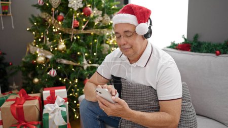 Foto de Hombre de mediana edad con el pelo gris usando el teléfono escuchando música por la decoración de Navidad en casa - Imagen libre de derechos