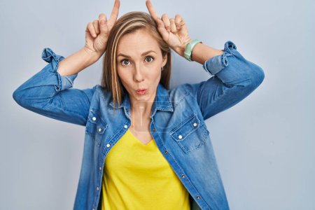 Photo for Young blonde woman standing over blue background doing funny gesture with finger over head as bull horns - Royalty Free Image