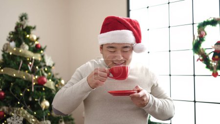 Photo for Young chinese man drinking coffee standing by christmas tree at home - Royalty Free Image