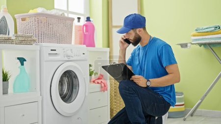Foto de Joven técnico hispano reparando lavadora hablando en smartphone en la lavandería - Imagen libre de derechos