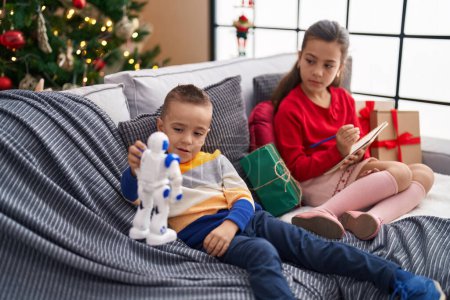 Foto de Hermano y hermana jugando con el robot y escribiendo en el cuaderno sentado en el sofá por el árbol de Navidad en casa - Imagen libre de derechos