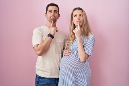 Téléchargez les photos : Jeune couple s'attendant à un bébé debout sur fond rose pensant concentré sur le doute avec le doigt sur le menton et regardant vers le haut se demandant - en image libre de droit