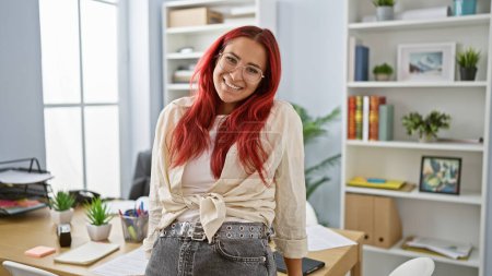 Téléchargez les photos : Une jeune femme d'affaires rousse confiante et souriante, une employée de bureau amusante et prospère assise à sa table de travail heureuse - en image libre de droit