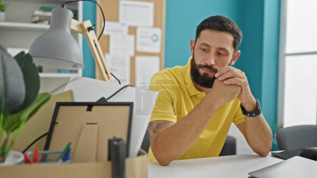 Photo for Young hispanic man being fired sitting on desk looking belongings package at the office - Royalty Free Image