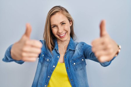 Téléchargez les photos : Jeune femme blonde debout sur fond bleu approuvant faire un geste positif avec la main, pouces levés souriant et heureux pour le succès. geste gagnant. - en image libre de droit