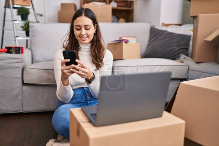 Photo for Young beautiful hispanic woman using laptop and smartphone sitting on floor at new home - Royalty Free Image