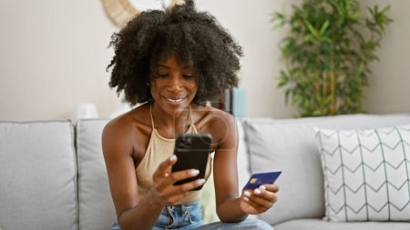 Foto de Mujer afroamericana de compras con teléfono inteligente y tarjeta de crédito sentado en el sofá en casa - Imagen libre de derechos