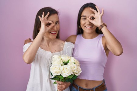 Téléchargez les photos : Mère et fille hispaniques tenant un bouquet de fleurs blanches faisant un geste correct avec la main souriante, les yeux regardant à travers les doigts avec un visage heureux. - en image libre de droit