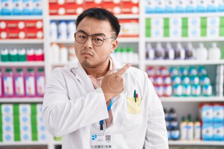 Foto de Joven chino trabajando en farmacia apuntando con el dedo de la mano a un lado mostrando publicidad, cara seria y tranquila - Imagen libre de derechos