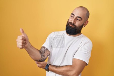 Téléchargez les photos : Jeune homme hispanique avec barbe et tatouages debout sur fond jaune regardant fier, souriant en faisant les pouces vers le haut geste sur le côté - en image libre de droit