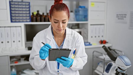 Photo for Beautiful, serious young redhead woman, the irish scientist hard at work at her lab table, writing research notes on her touchpad amidst the hustle of technology-filled, bustling medical laboratory. - Royalty Free Image