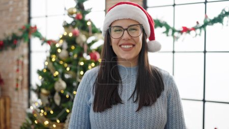 Foto de Joven mujer hispana sonriendo de pie junto al árbol de Navidad en casa - Imagen libre de derechos