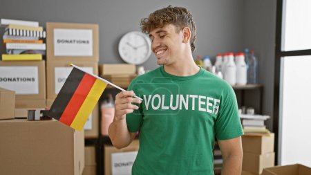 Téléchargez les photos : Portrait chaleureux, jeune homme hispanique souriant, bénévole dévoué dans un centre caritatif, tenant fièrement le drapeau allemand - en image libre de droit