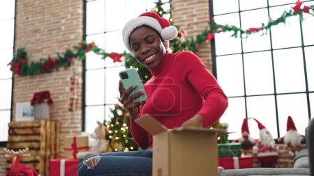 Foto de Mujer afroamericana usando teléfono inteligente desembalaje regalo de Navidad en casa - Imagen libre de derechos