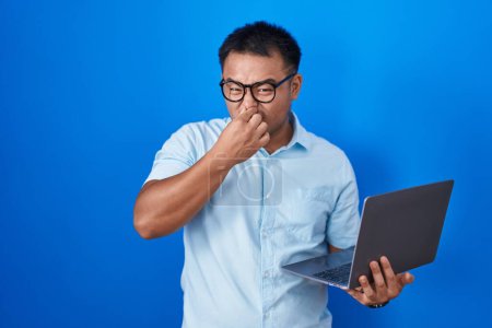 Téléchargez les photos : Jeune homme chinois utilisant un ordinateur portable sentant quelque chose de puant et dégoûtant, odeur intolérable, retenant souffle avec les doigts sur le nez. mauvaise odeur - en image libre de droit