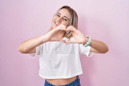 Photo for Young blonde woman standing over pink background smiling in love doing heart symbol shape with hands. romantic concept. - Royalty Free Image