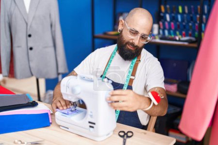 Foto de Young bald man tailor smiling confident using sewing machine at clothing factory - Imagen libre de derechos