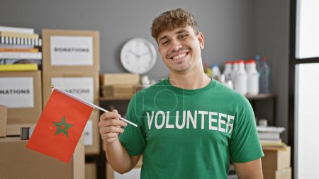 Photo for Radiant young hispanic man, proudly holding moroccan flag, contributes as smiling volunteer at vibrant community charity center - Royalty Free Image